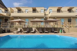 Una piscina con sillones de descanso y sombrillas, rodeada por un edificio con balcones y un muro de piedra.