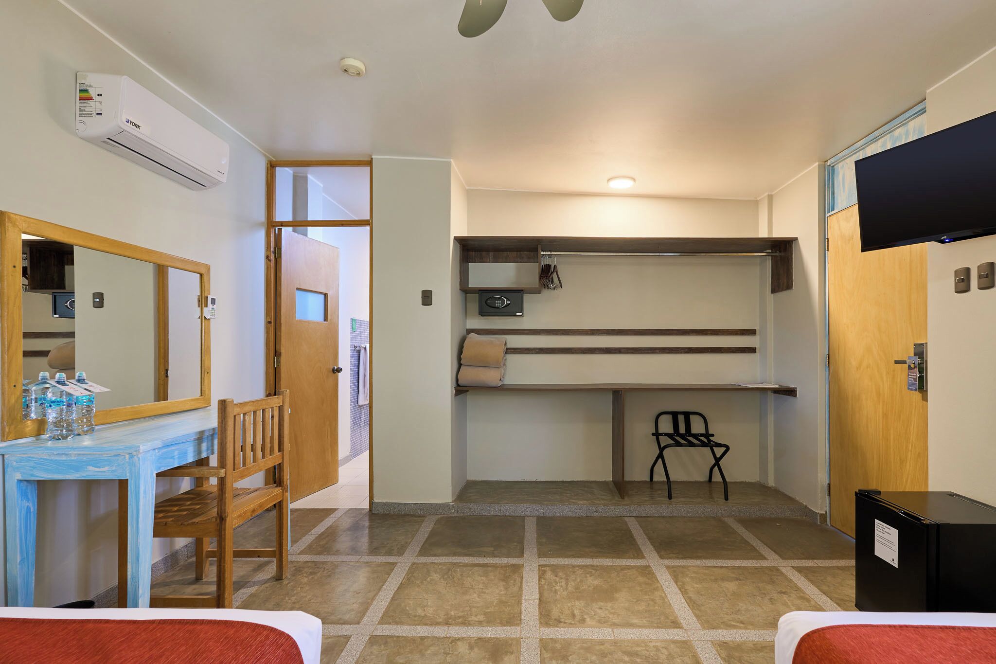 Habitación pequeña con cama, escritorio con silla, una mesita con botellas, espejo, estante con silla y televisión.