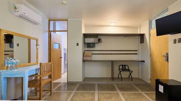 Habitación pequeña con cama, escritorio con silla, una mesita con botellas, espejo, estante con silla y televisión.