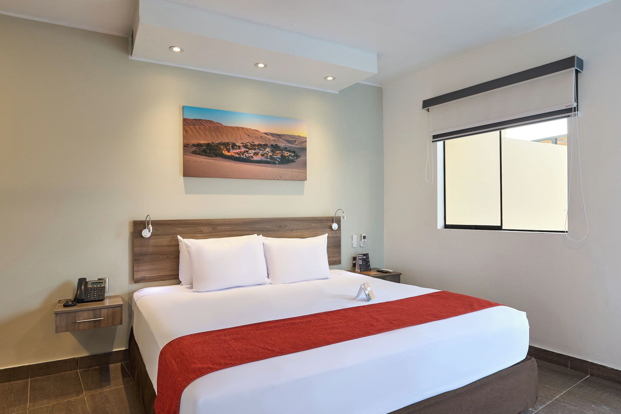 Une chambre d’hôtel avec un grand lit, un tableau représentant un paysage désertique, une fenêtre avec des stores et un téléphone sur la table de chevet.