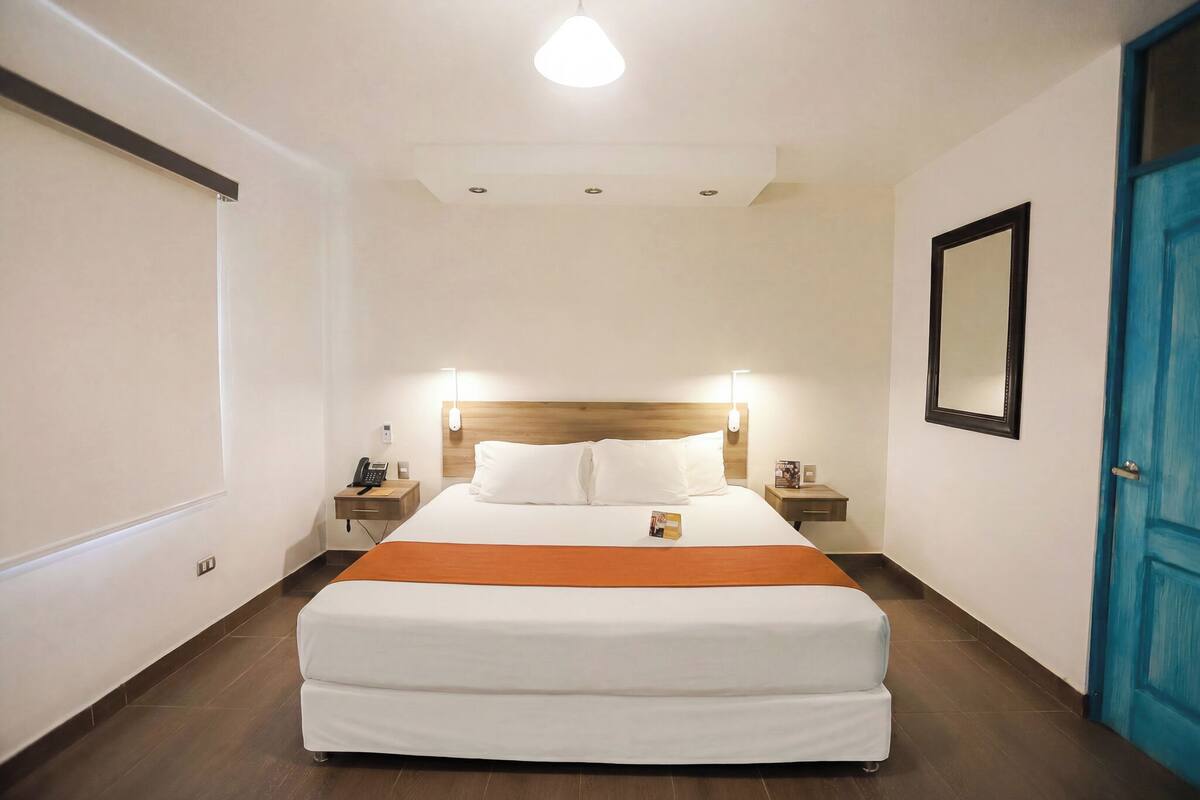 A hotel room with a bed, bedside tables, a nightstand, a mirror, and a blue door.