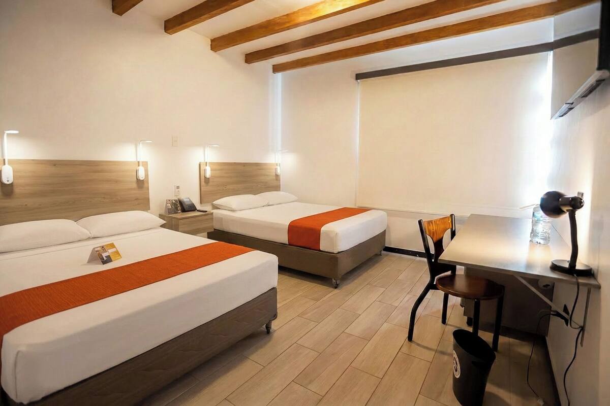 A hotel room with two beds, a desk, a chair, a trash bin, and a window.