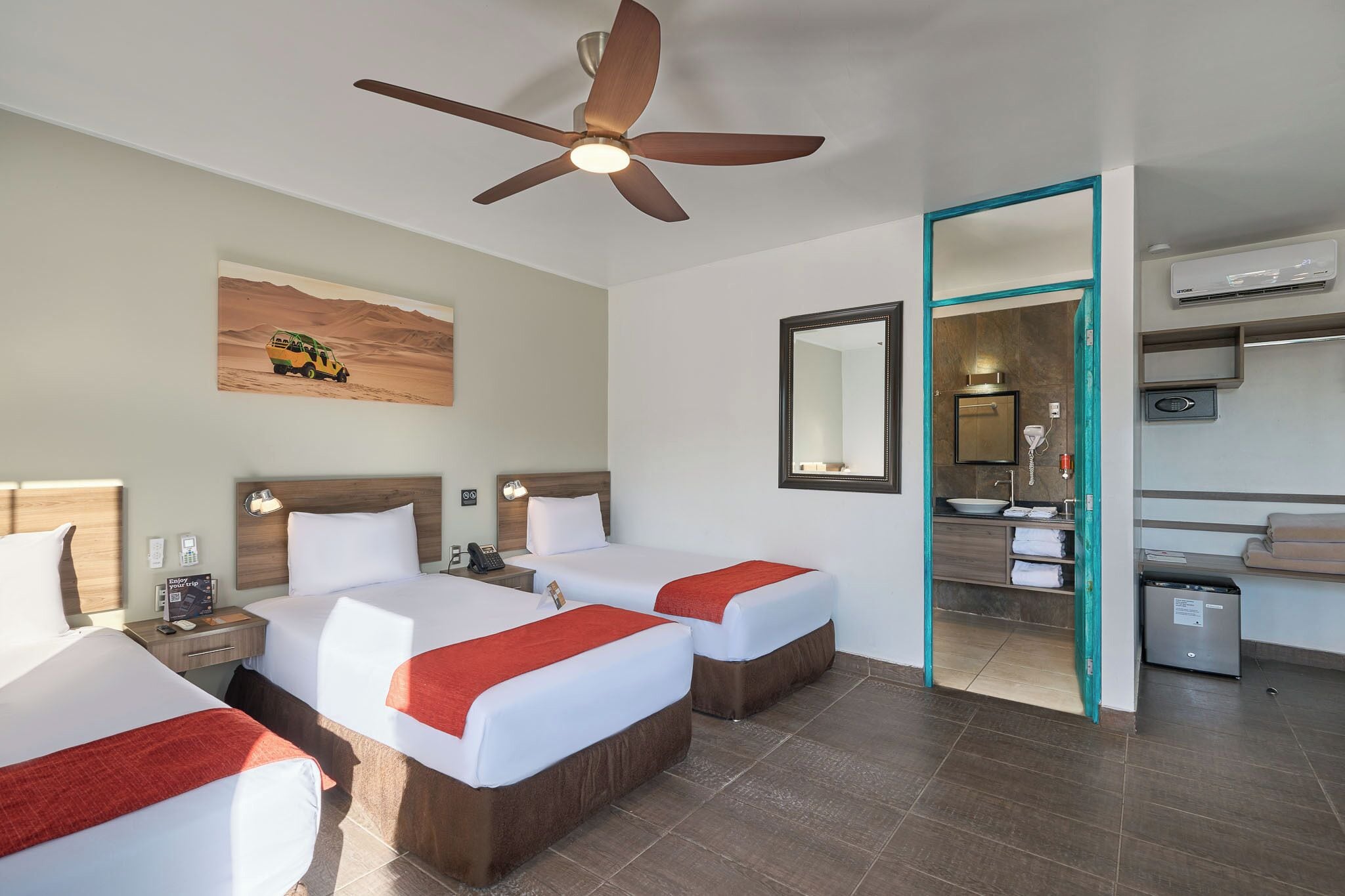 Habitación de hotel con dos camas, un ventilador de techo, un cuadro, un espejo, un baño con lavamanos y un equipo de aire acondicionado.