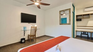 Habitación de hotel con una cama, ventilador de techo, televisor montado, una silla, una mesita con botellas de agua y un espejo.
