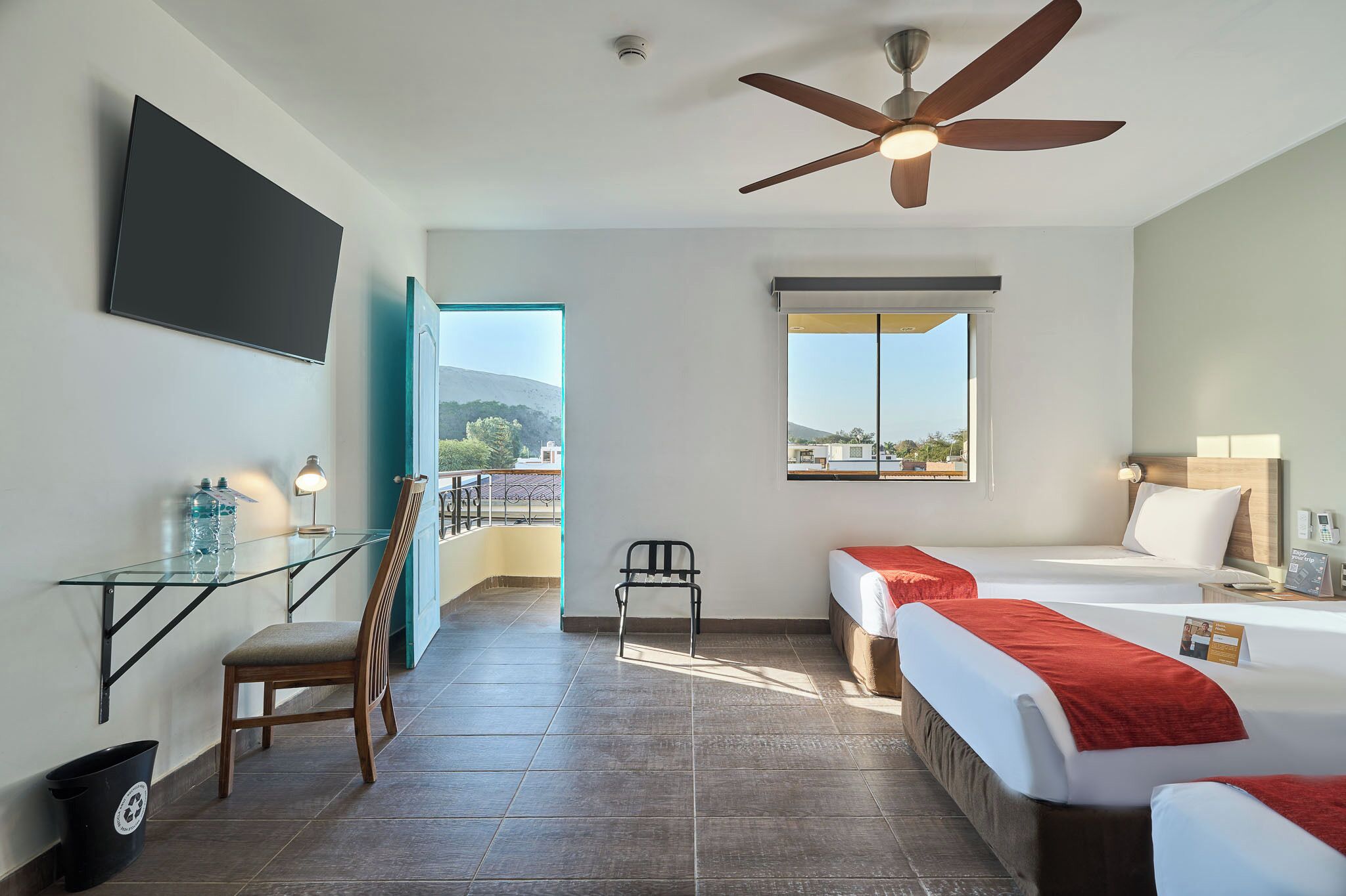 Habitación de hotel con cama, escritorio, silla, televisor montado en la pared y ventilador de techo.