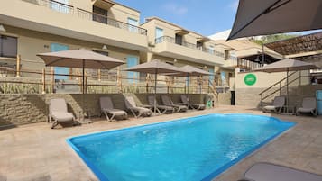 A swimming pool with lounge chairs and umbrellas, a stone wall, and a building with balconies.