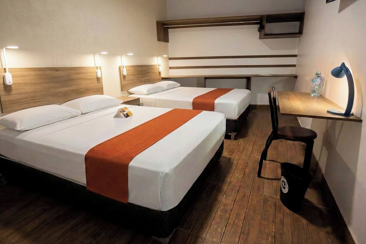A hotel room with two beds, a desk, a chair, and a trash bin.