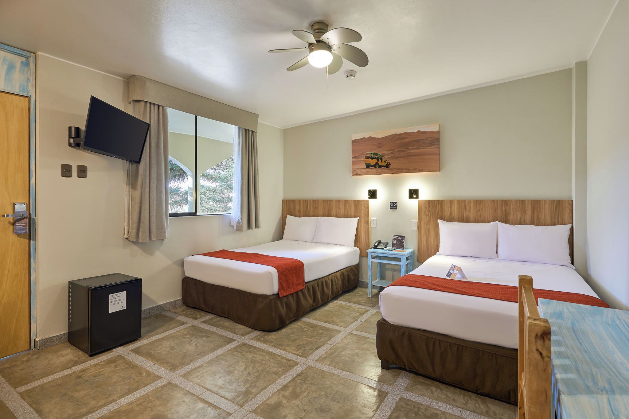 Une chambre d’hôtel avec deux lits, une télévision, un ventilateur de plafond, une fenêtre avec des rideaux, une petite table et un réfrigérateur.