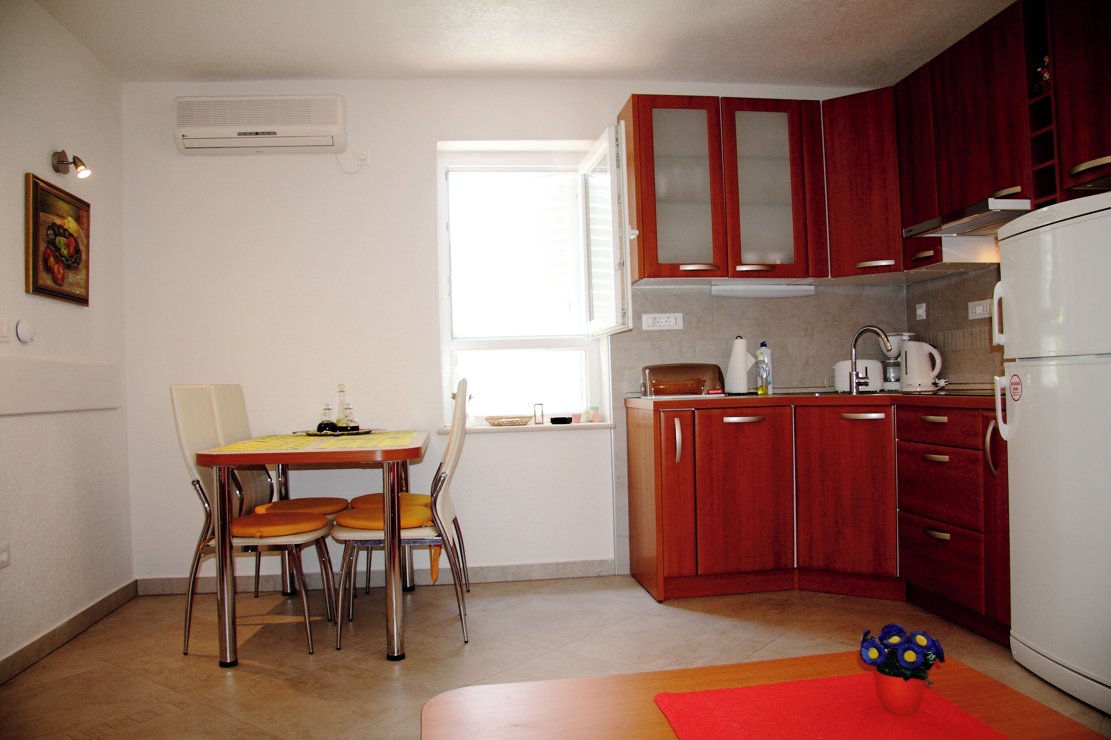 A compact kitchen with wooden cabinets, a white refrigerator, a dining table with chairs, and a window providing natural light.