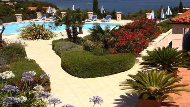 Zona de piscina con sillones de descanso y sombrillas, rodeada de jardines ajardinados y vistas al mar y a las montañas.
