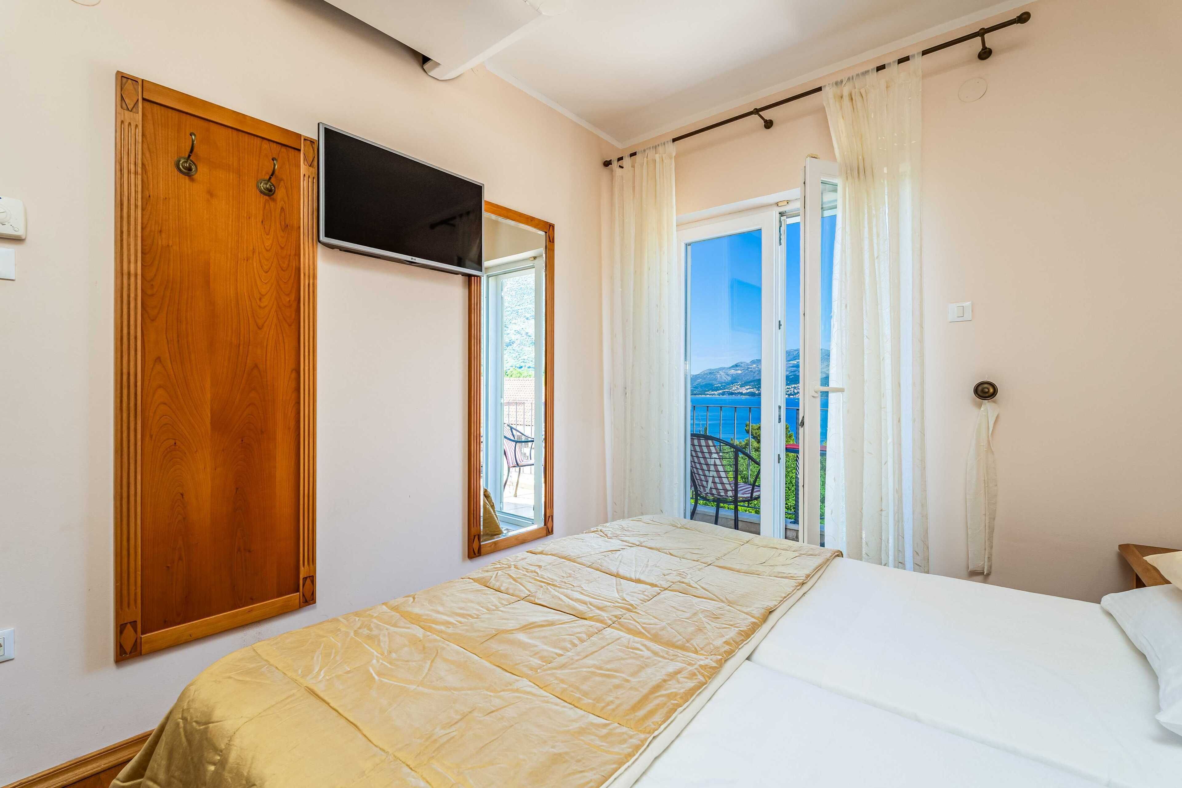 A bedroom with a bed, a TV mounted on the wall, a wooden wardrobe, and a balcony with a view.