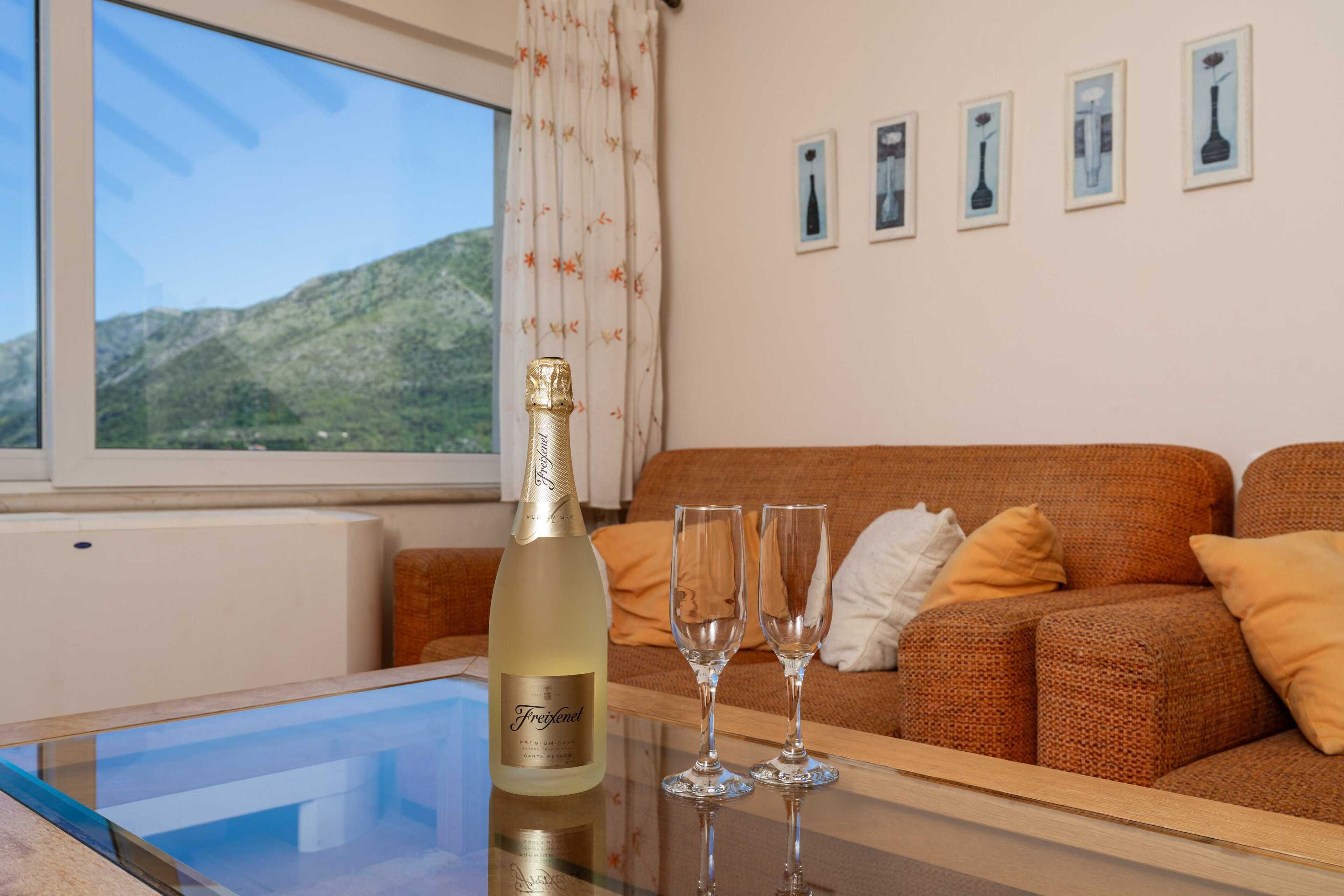 A room with a glass table, a bottle of champagne, and two glasses on it, a brown sofa with cushions, and a window with a view of hills.