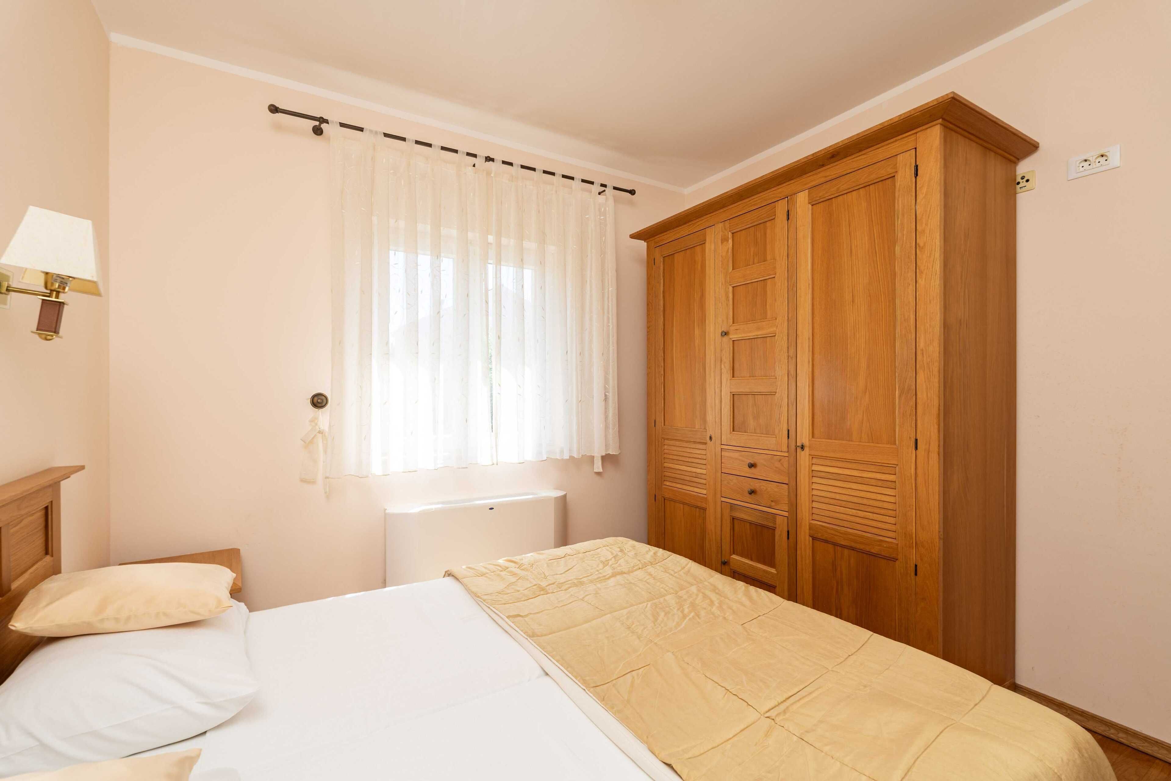 A bedroom with a bed, a wooden wardrobe, a window with sheer curtains, and a wall-mounted lamp.