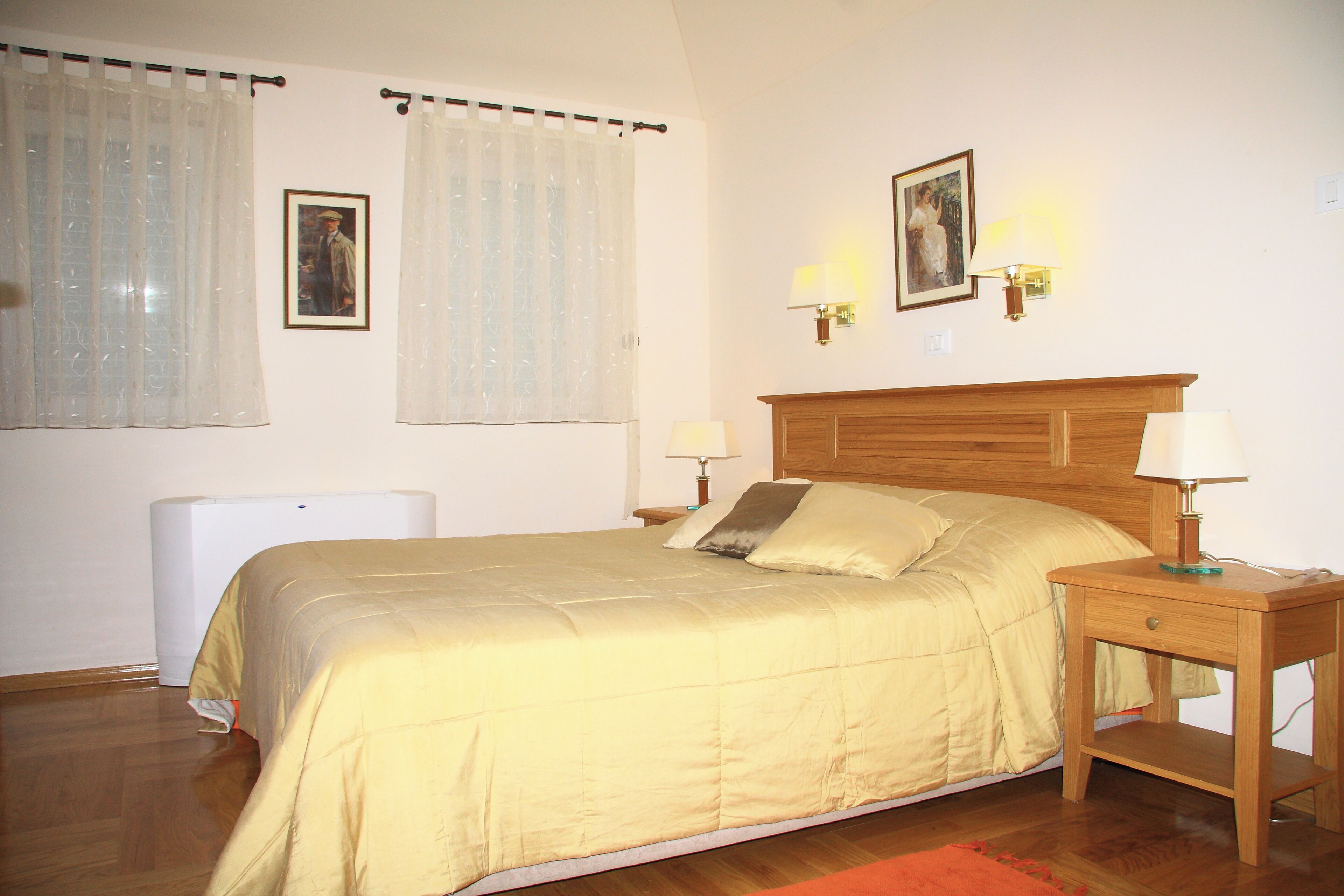 Quarto com cama, duas mesas de cabeceira com luminárias, um radiador e duas janelas com cortinas.