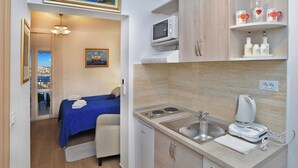Ein kleines Hotelzimmer mit einem Bett, einer kleinen Küchenzeile, einer Mikrowelle, einem Spülbecken und einer Kaffeemaschine. An der Wand hängt ein Gemälde und durch das Fenster hat man Blick auf das Meer.