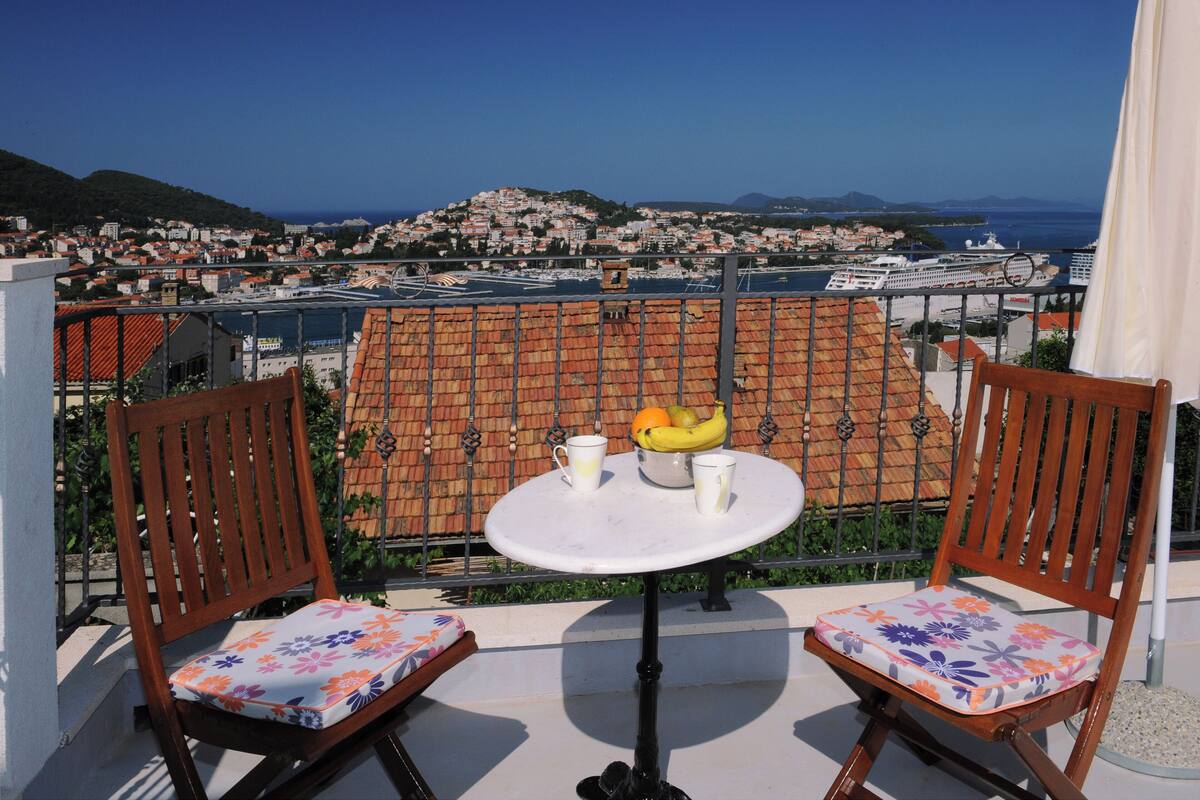 Un balcon avec deux chaises en bois et une petite table, d’où l’on peut admirer une ville côtière avec ses bâtiments et un navire de croisière au loin.