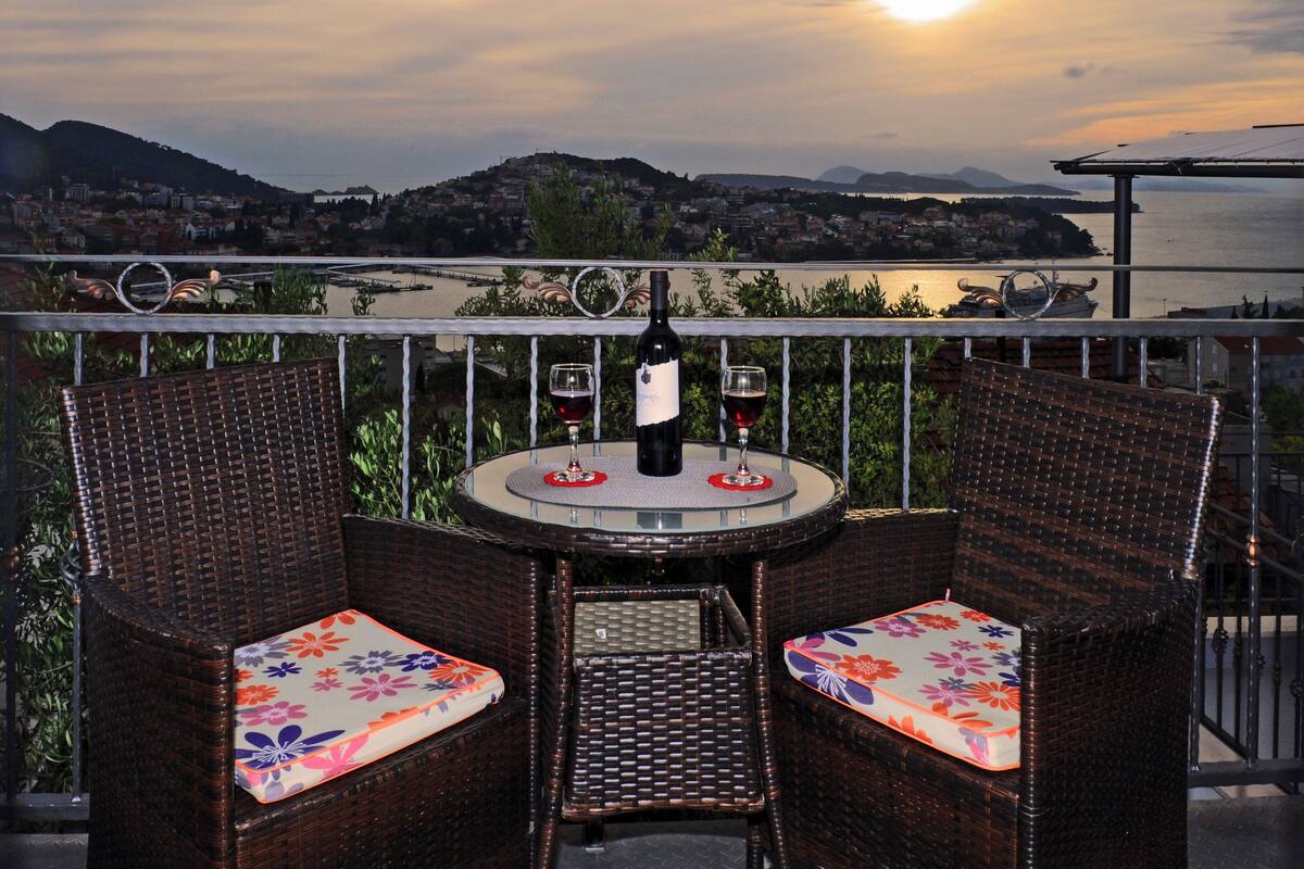 Un balcon agrémenté de meubles en osier, une table avec une bouteille et des verres, et une vue panoramique sur une ville et des montagnes.