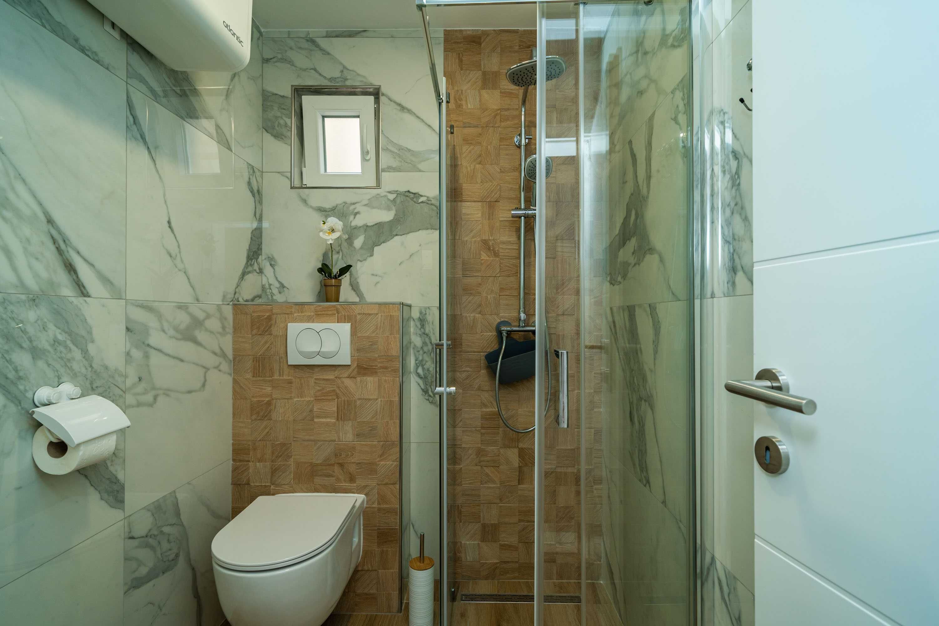 A bathroom with a toilet, a shower with a glass door, marble walls, and a wooden panel accent.