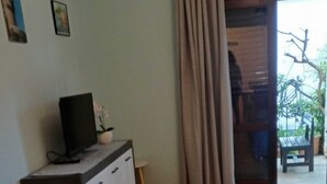 Quarto com piso de madeira, sofá, cama, TV sobre um móvel e vista do exterior através de uma janela com cortinas.