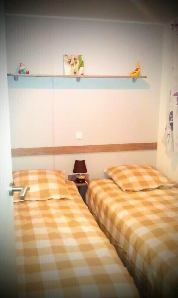 A bedroom with two beds, each with a checkered bedspread, a nightstand with a lamp, and a shelf above the beds. - Camping Les Truffieres (Saint-Marcel-d'Aredeche)