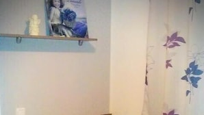 Een slaapkamer met een geruite sprei, een nachtkastje met een lamp, een plank met een ingelijst foto en een bloemenprint gordijn.