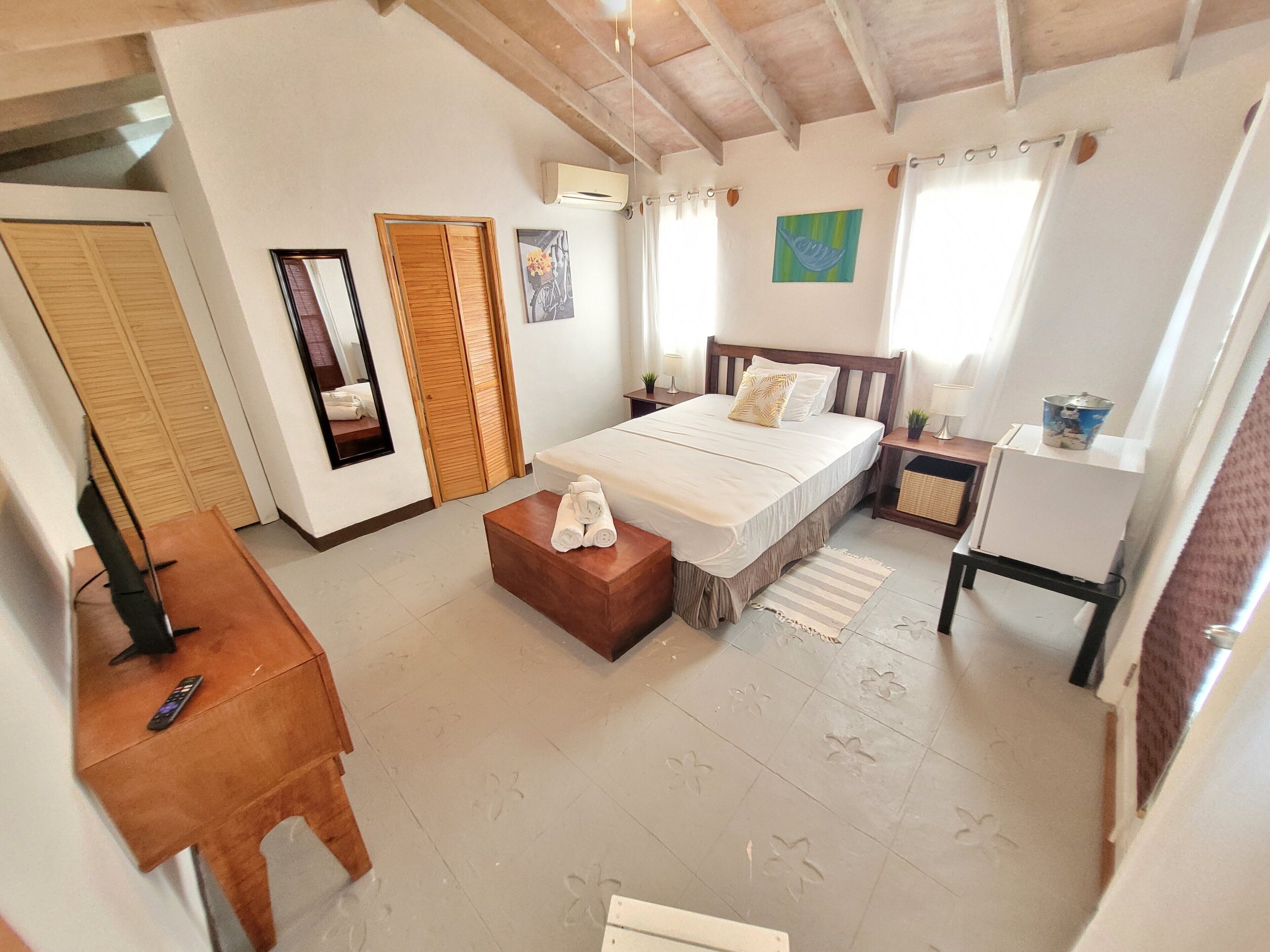 A bedroom with a bed, a wooden nightstand, a small table with towels, a mirror, and a window with curtains.