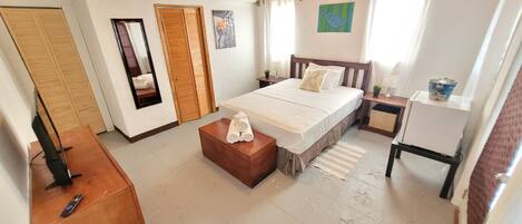 A bedroom with a bed, a wooden nightstand, a small table with towels, a mirror, and a window with curtains.