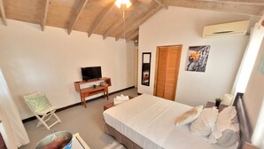 Quarto com cama, televisão sobre suporte de madeira, cadeira, mesa pequena e um balde.