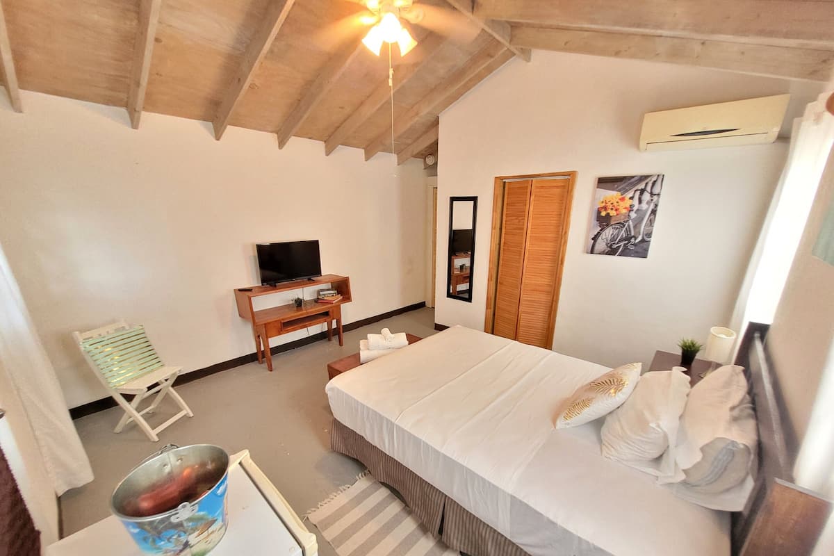 A bedroom with a bed, a TV on a wooden stand, a chair, a small table, and a bucket.