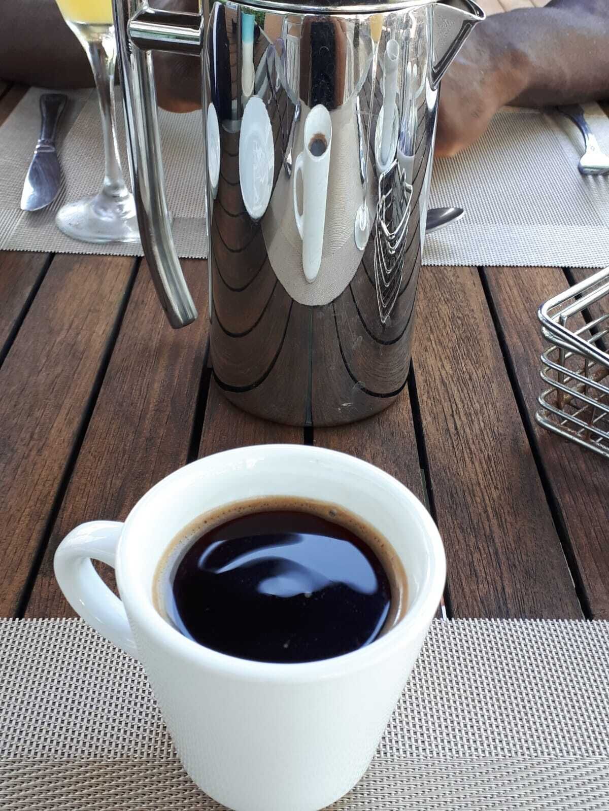 Een witte koffiekop op een houten tafel, met een metalen kan op de achtergrond.