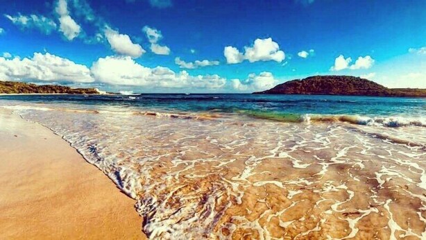Uma praia com águas claras e rasas, uma faixa de areia e uma ilha ao longe.