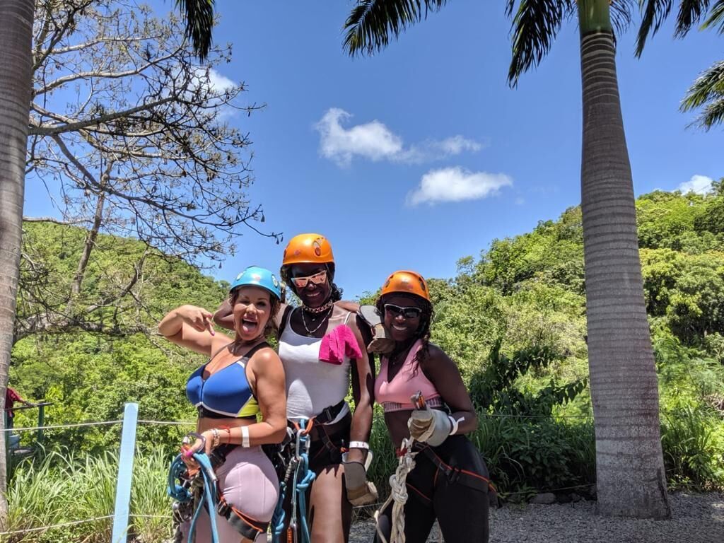 A group of people on a zip-lining adventure, wearing helmets and harnesses, with a natural landscape and palm trees in the background.