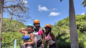 A group of people on a zip-lining adventure, wearing helmets and harnesses, with a natural landscape and palm trees in the background.
