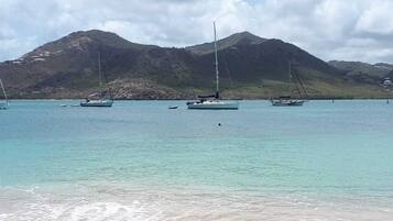 Una playa con varios veleros anclados en aguas turquesas tranquilas, una orilla arenosa y un telón de fondo montañoso.