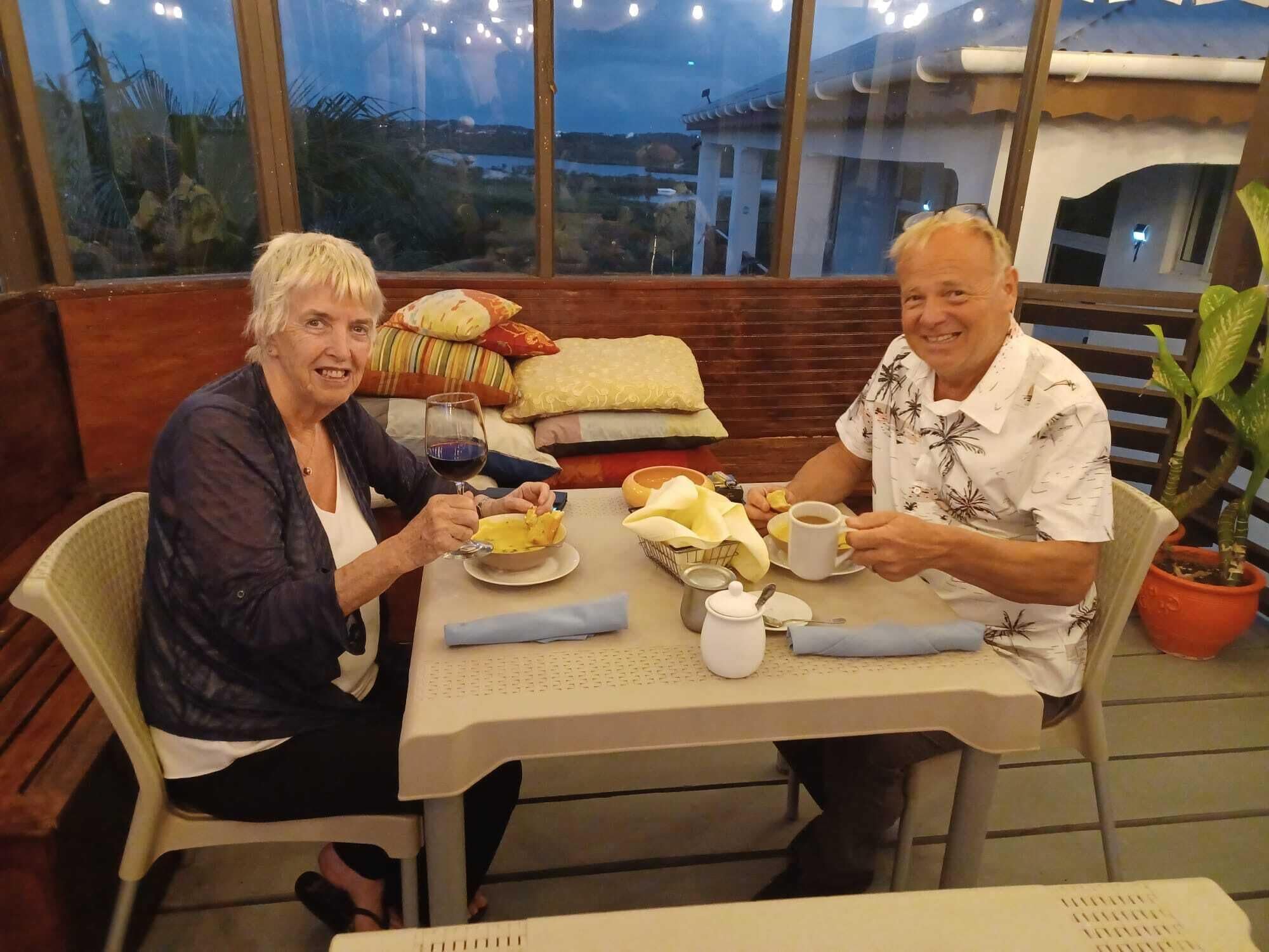 Two people seated at a table with a view of the outdoors. The table has a white tablecloth, a basket, a cup, and a plate with food.