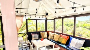Uma área de estar coberta ao ar livre com mesa e cadeiras, janelas amplas com vista para o verde e luzes de fada.