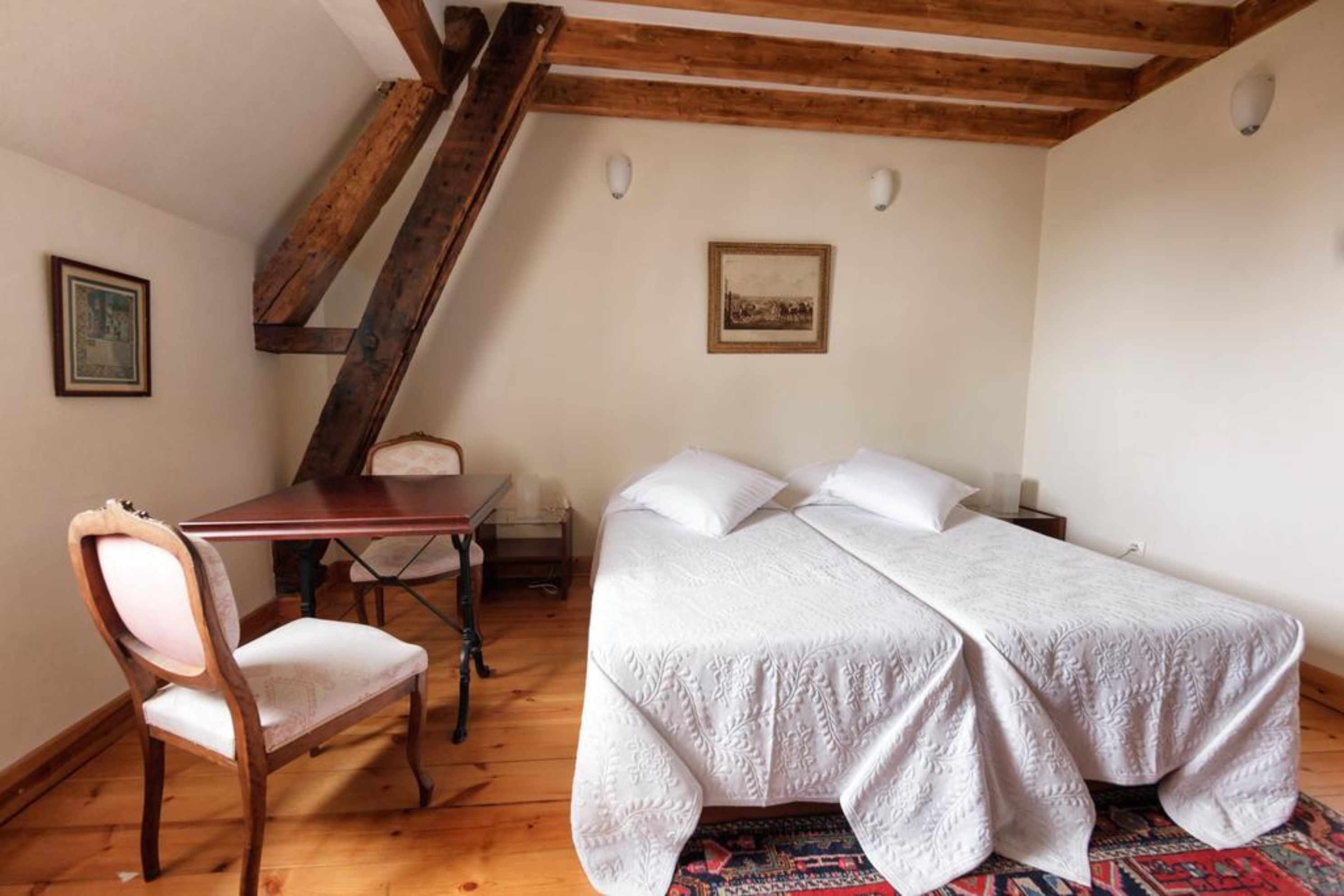 A bedroom with a wooden ceiling, a bed with white bedding, a wooden chair, a small table, and two wall-mounted lights.