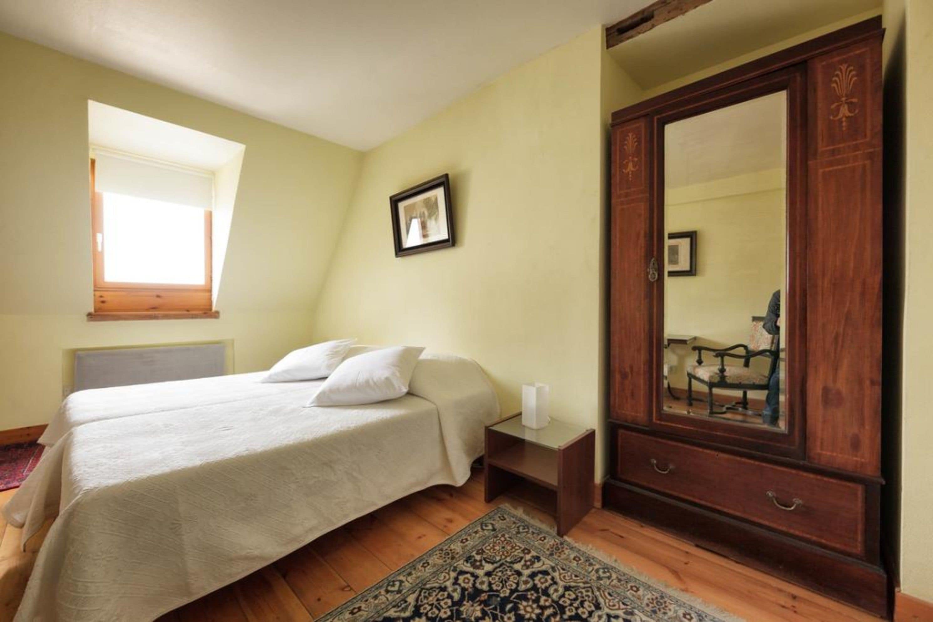 Een slaapkamer met een bed, een houten garderobe, een nachtkastje, een lijst met een foto aan de muur en een raam met uitzicht.