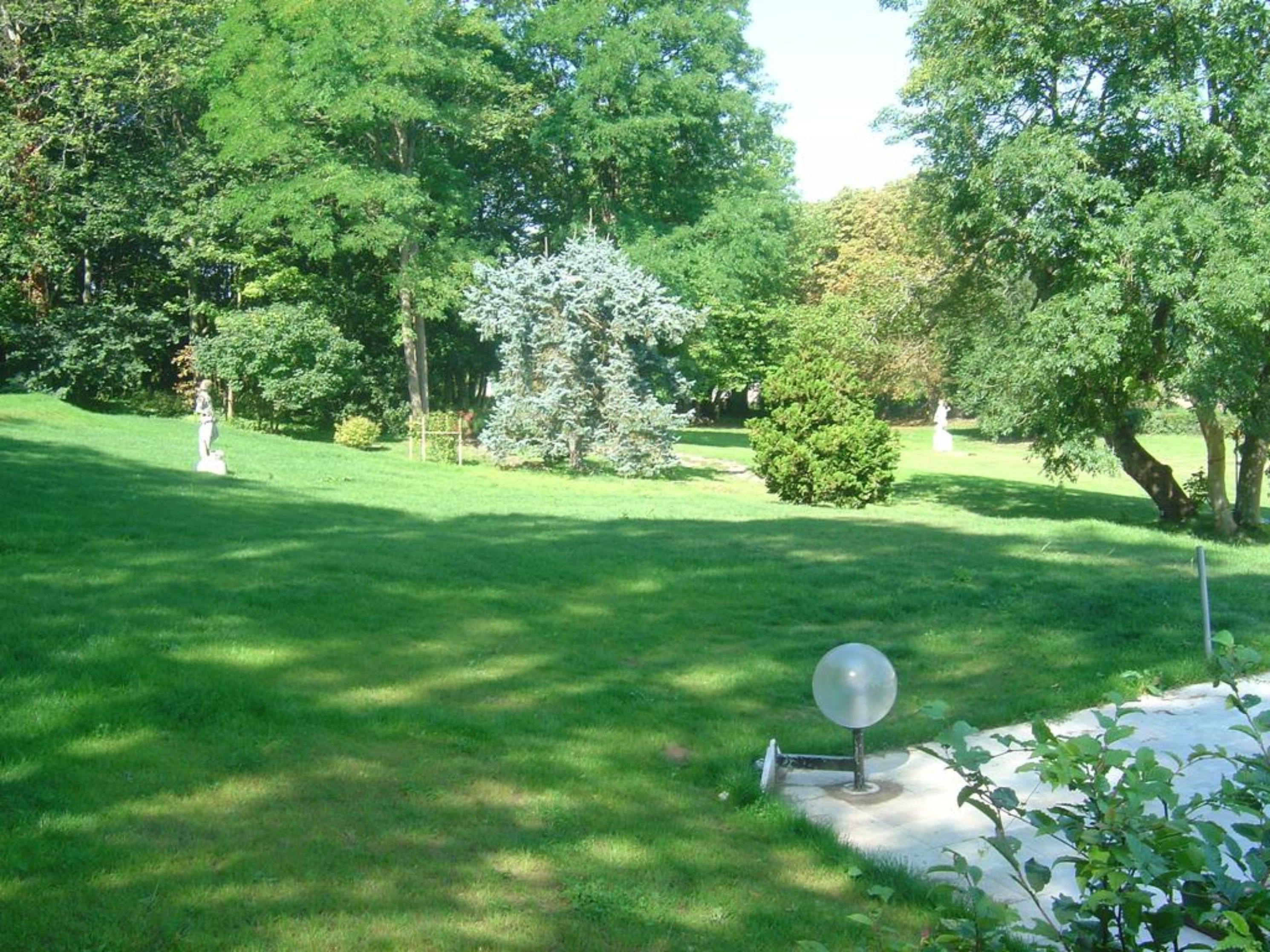 A well-maintained grassy area with a spherical sculpture, surrounded by trees and a statue in the background.