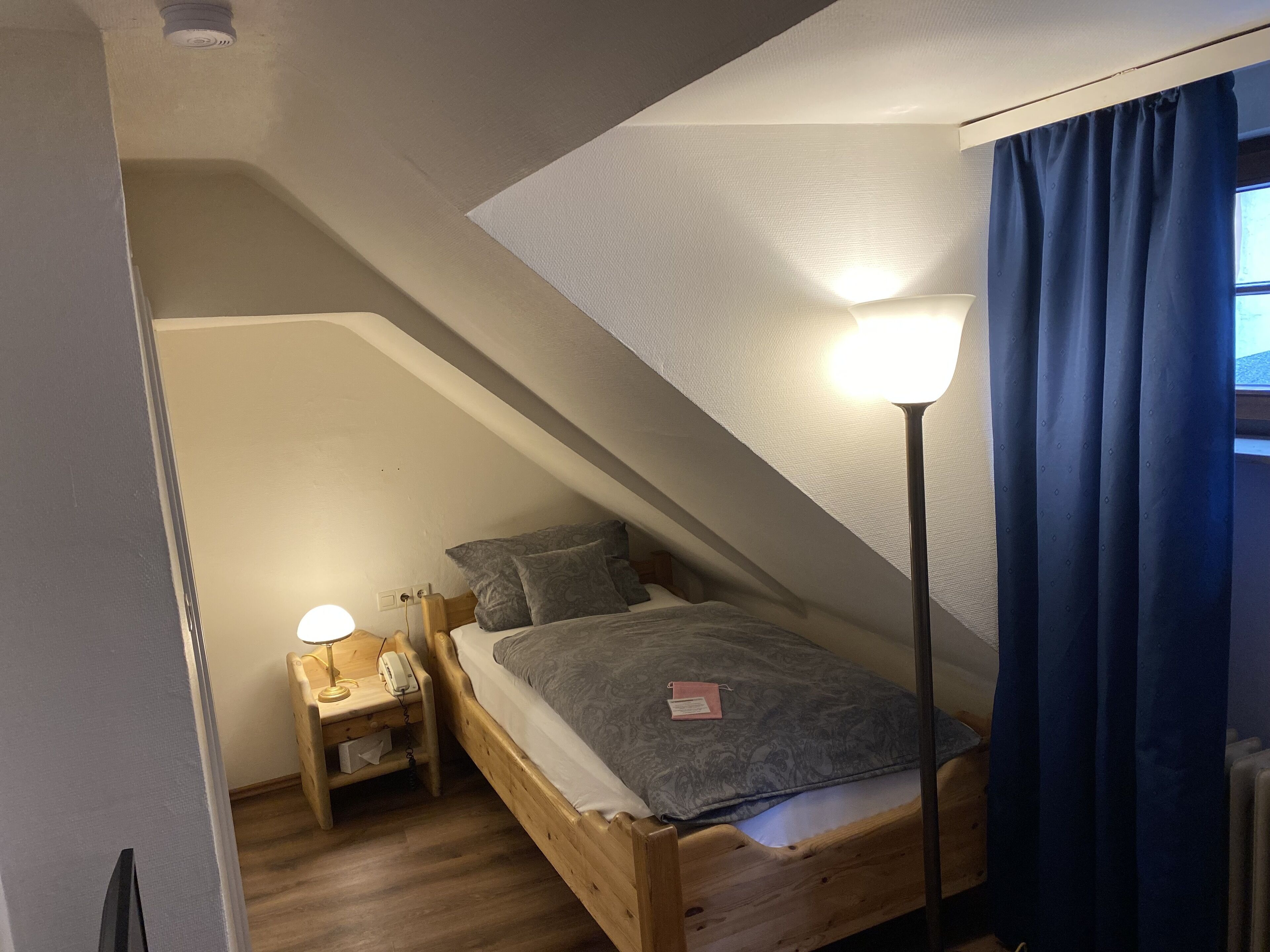 Ein kleines, gemütliches Schlafzimmer im Dachgeschoss mit schrägen Wänden, einem hölzernen Bett, einem Nachttisch mit Lampe und einem Fenster mit blauen Vorhängen.