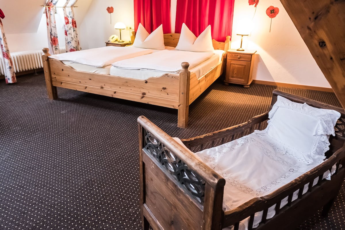 A wooden bed with two pillows and a nightstand with a lamp in a room with a sloped ceiling and red curtains.