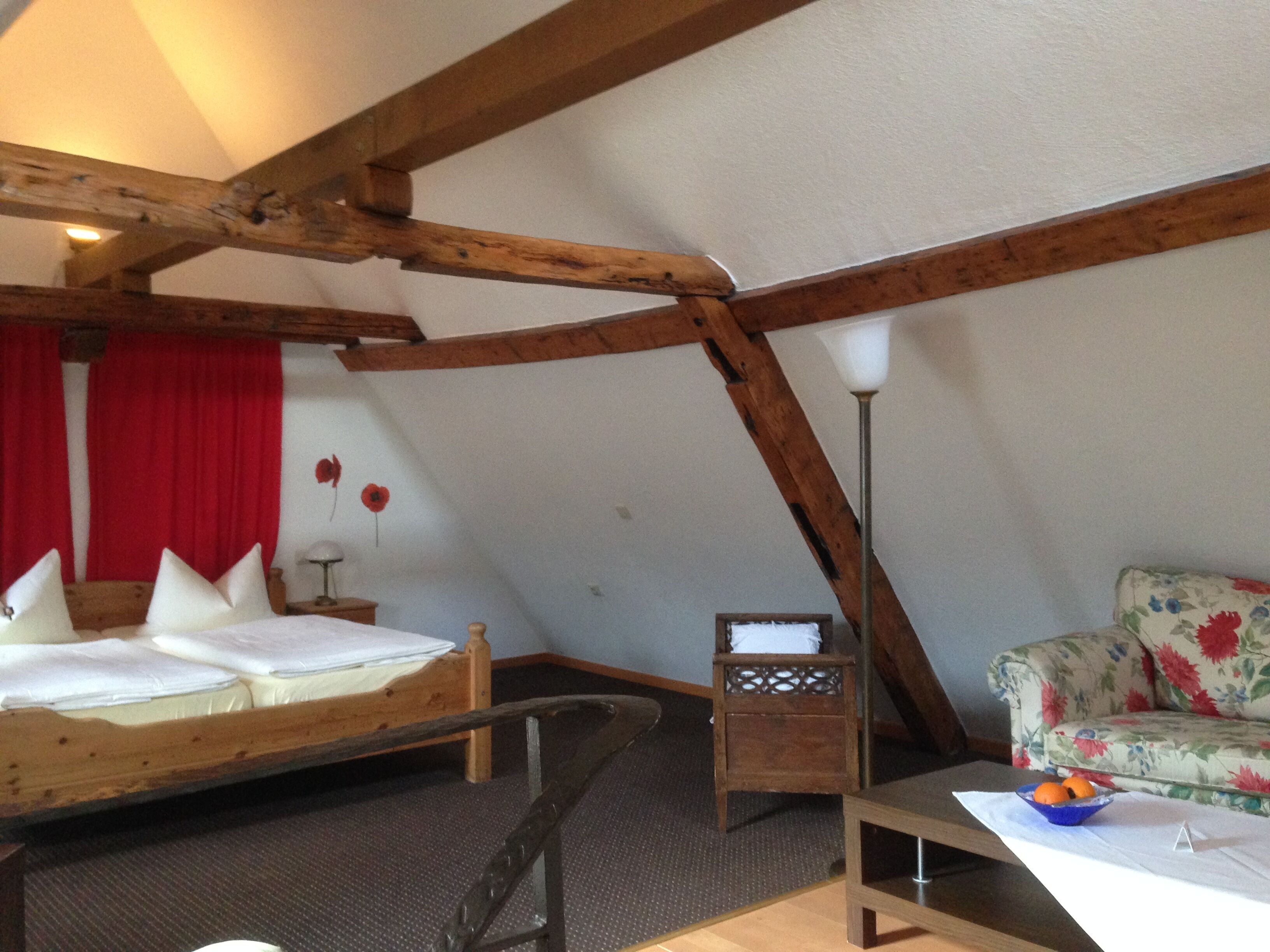 A cozy attic bedroom with a wooden ceiling, a bed with pillows, a small table with a bowl of fruit, and a floral sofa.