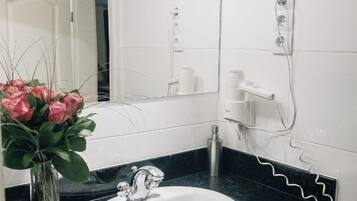 Een badkamer met een witte wastafel, een zwart aanrecht en een spiegel erboven. Op het aanrecht staat een vaas met roze bloemen.