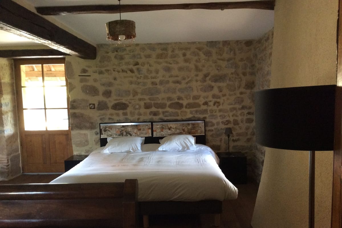 A bedroom with a stone wall, a wooden bed frame, a bedside table with a lamp, and a window with wooden shutters.
