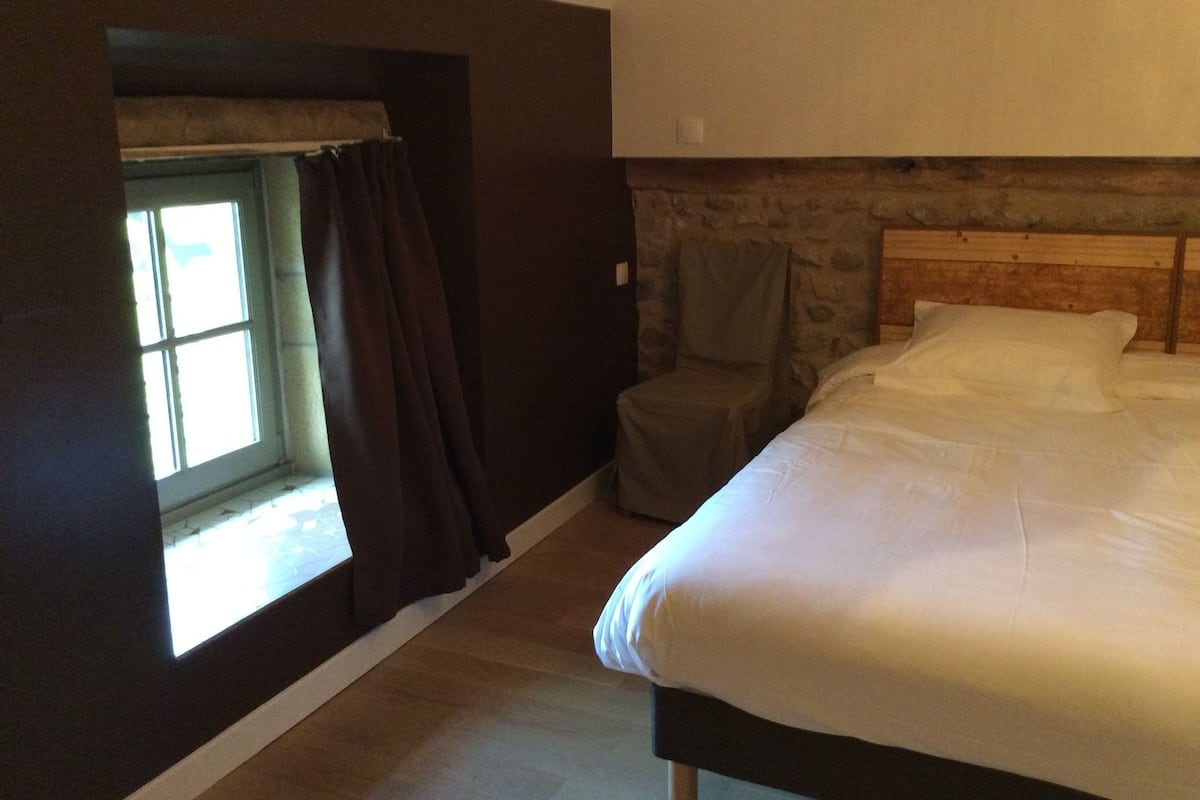 A bedroom with a large bed, a window with curtains, a stone wall, and a chair.