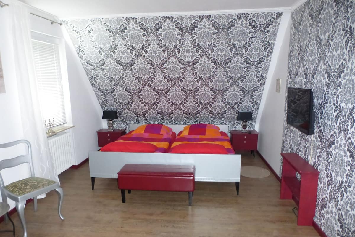 Une chambre avec un papier peint à motifs, un lit avec une literie colorée, deux tables de chevet, un banc rouge, une chaise et un téléviseur fixé au mur.
