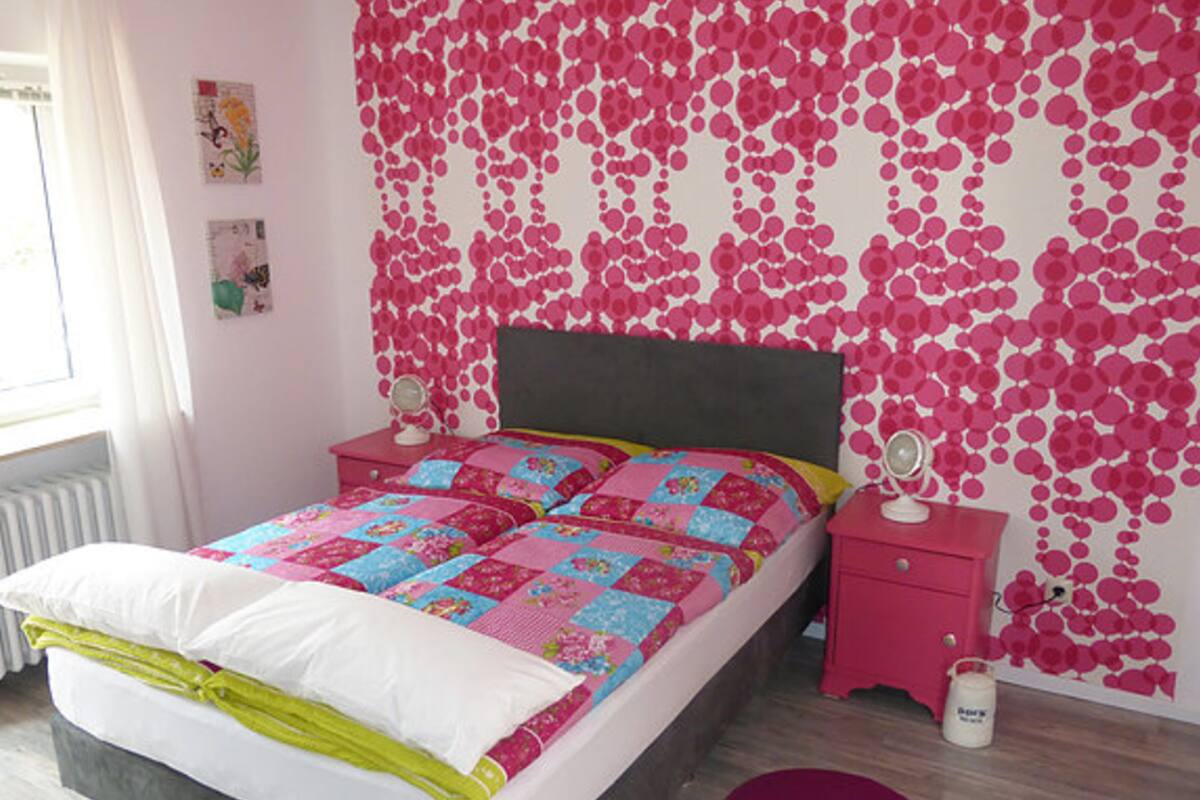 Une chambre à coucher avec un lit, des tables de chevet roses et un mur orné d’un motif rose et blanc.