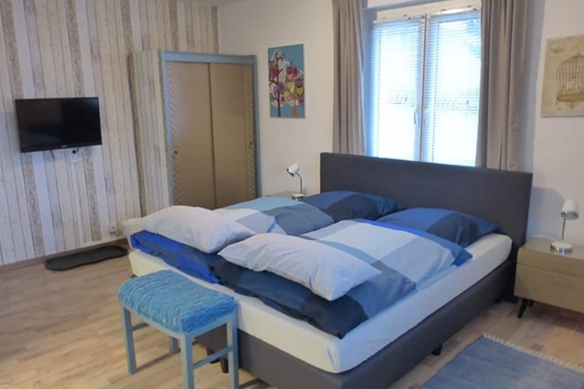 Une chambre à coucher avec un lit, un banc, une télévision, une armoire et une fenêtre avec des rideaux.