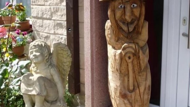 A wooden gnome statue with a hat, a stone angel statue, and potted plants in front of a building with a stone facade and a door.