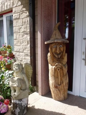 Een houten tuinbeeldje van een kabouter, een stenen engelbeeld en potplanten voor een gebouw met een stenen gevel en een deur.